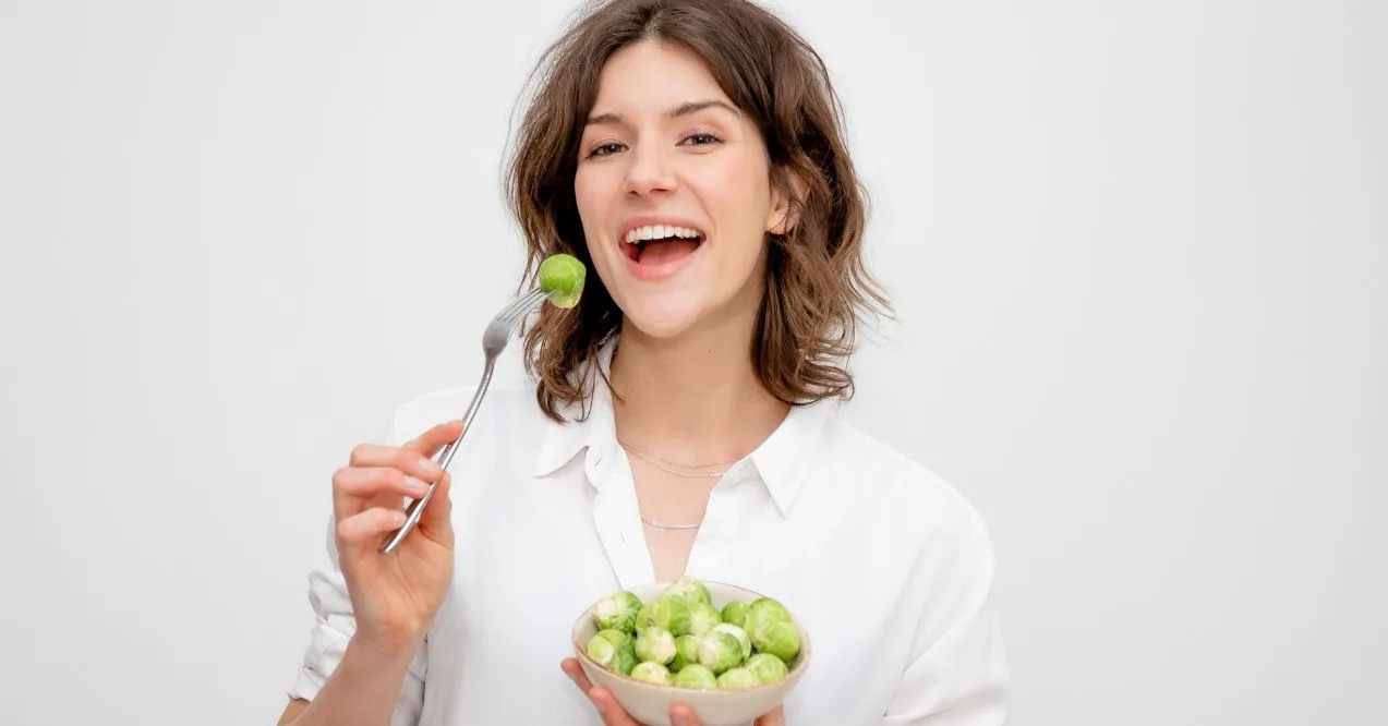 Woman in a white shirt holding a fork with a Brussels sprout and a bowl of sprouts in her other hand.