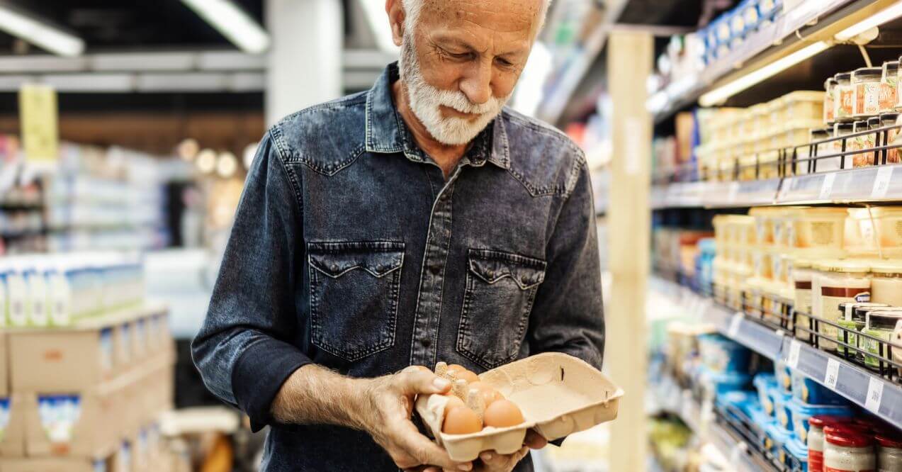 Older man holding carton of eggs in supermarket aisle.