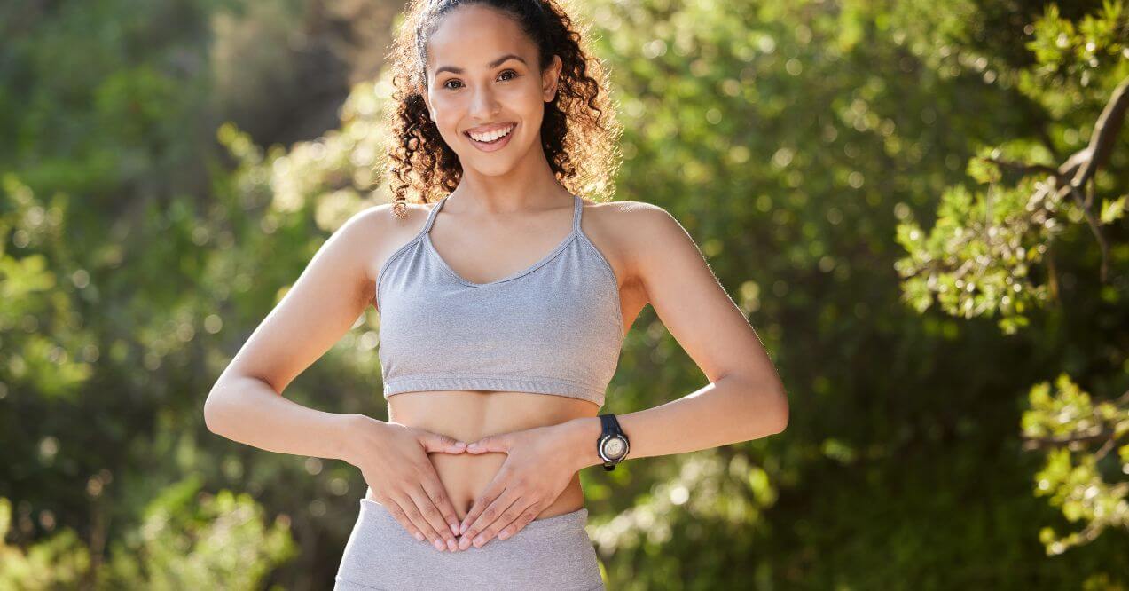 Smiling woman outdoors in workout clothes forming heart shape over stomach, promoting gut health program.