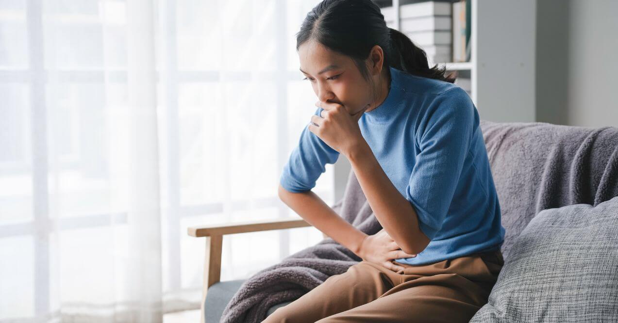 Woman sitting on couch feeling pain in abdomen while coughing.