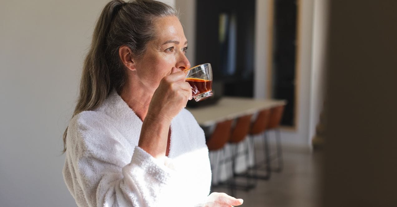 Woman in robe drinking tea in a bright kitchen wondering can green tea cause bloating