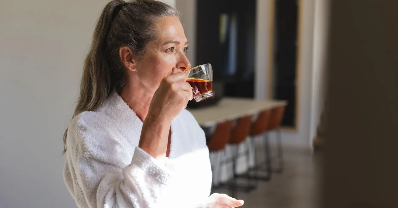 Woman in robe drinking tea in a bright kitchen wondering can green tea cause bloating